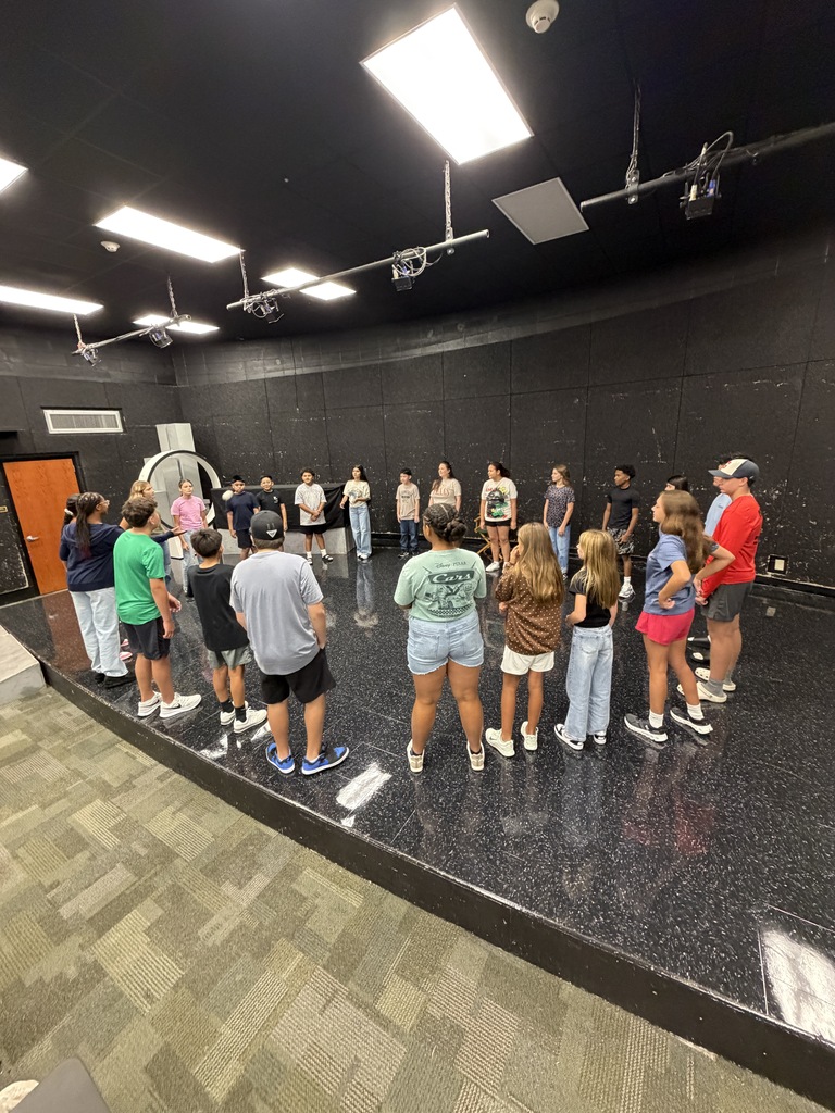 Several people stand in a circle on a stage in a room with black walls, ceiling, and tiled floor.