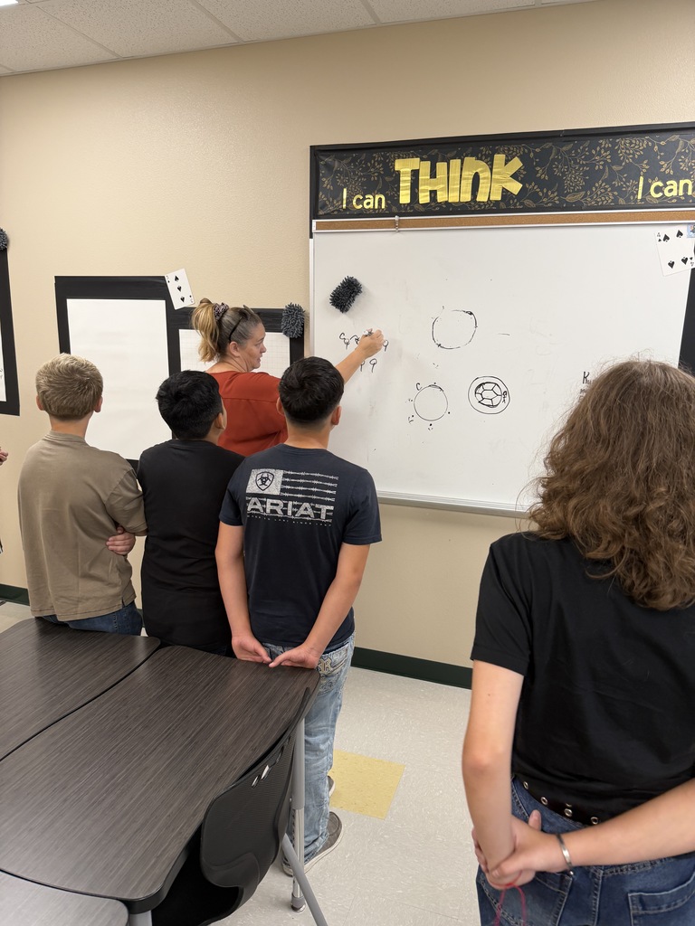 Five students look at a whiteboard displaying the word "THINK" and circles. They stand in a classroom with tables and beige walls.
