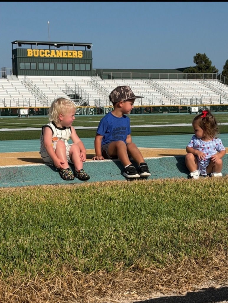 Three children sit on the edge of a field at a sports stadium, with a "Buccaneers" sign in the background.