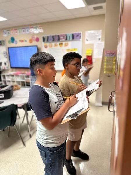 Two students with glasses stand in a classroom with papers, one holding a clipboard. Behind them, a monitor, tables, chairs, and bulletin board are visible.