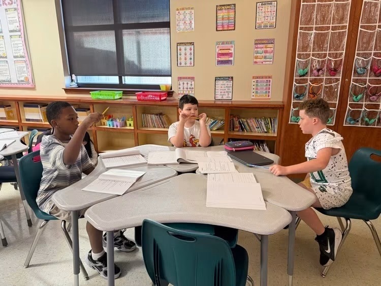Three boys seated at a table, one gesturing with a pencil, another reading, and the third writing. Shelves with books in the background.