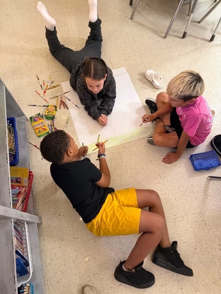 Three children in a classroom setting, each using colored pencils on a large paper. One lying down, two sitting.