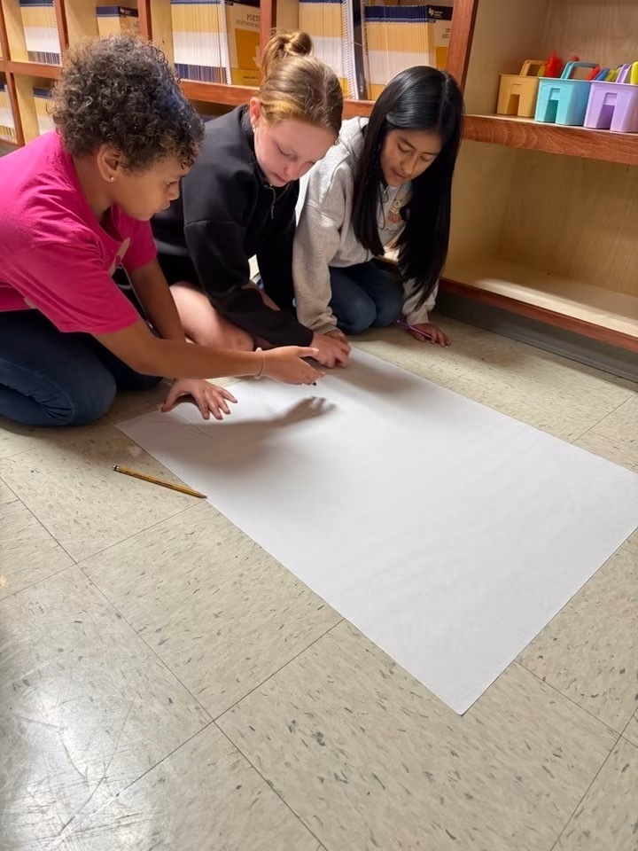 Three students work on a large white paper on the floor. A pencil rests on the paper.