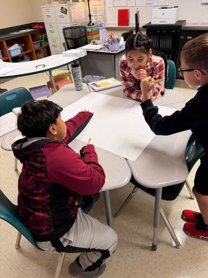 Three children sit around a table with papers and pencils in a classroom with desks and chairs.