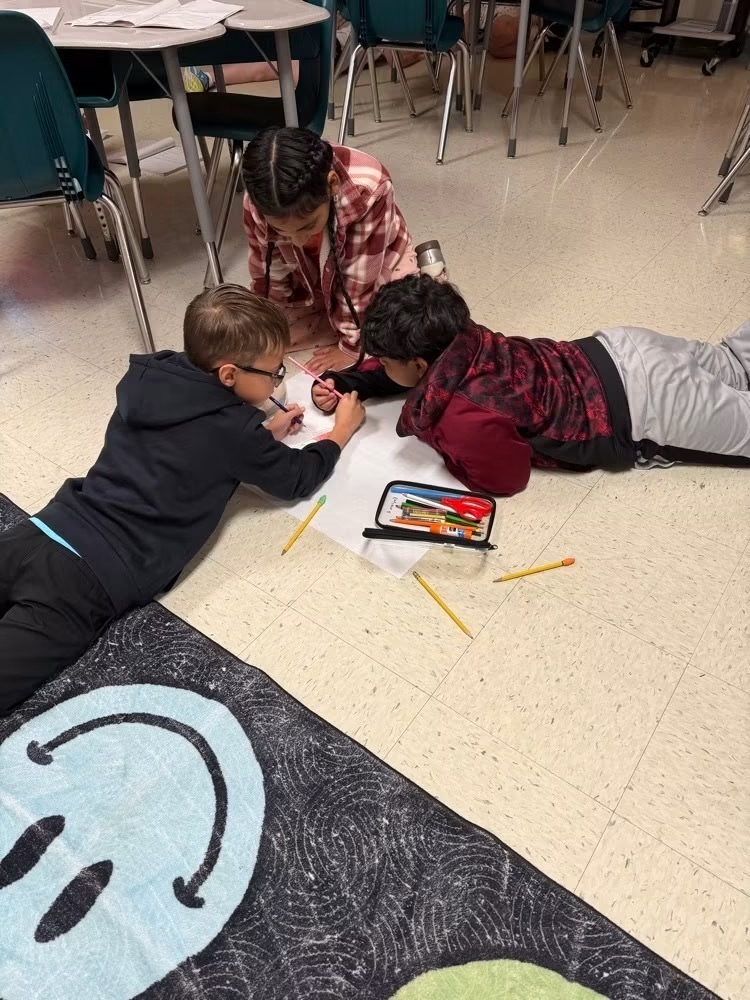 Three children are on the floor, drawing on paper. One child is seated, another lying down, and a third is on their side.