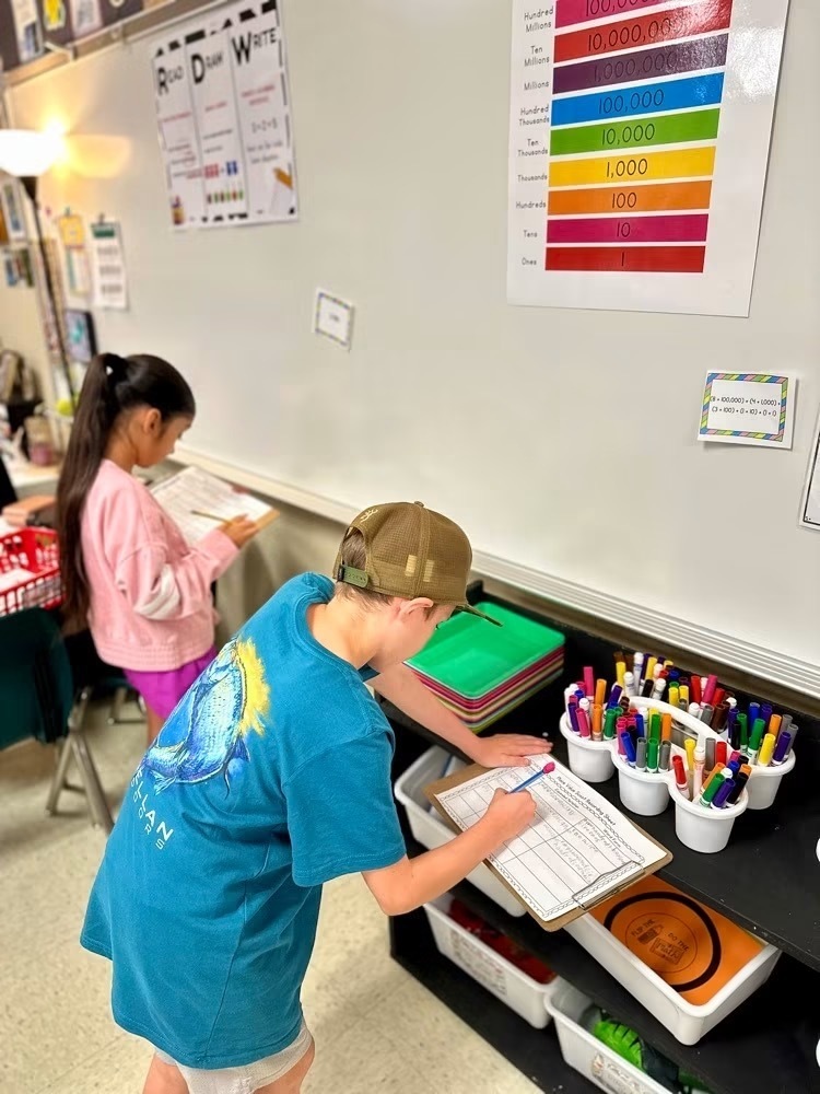 Children using markers and a clipboard at a table in a classroom with a colorful number chart on the wall.