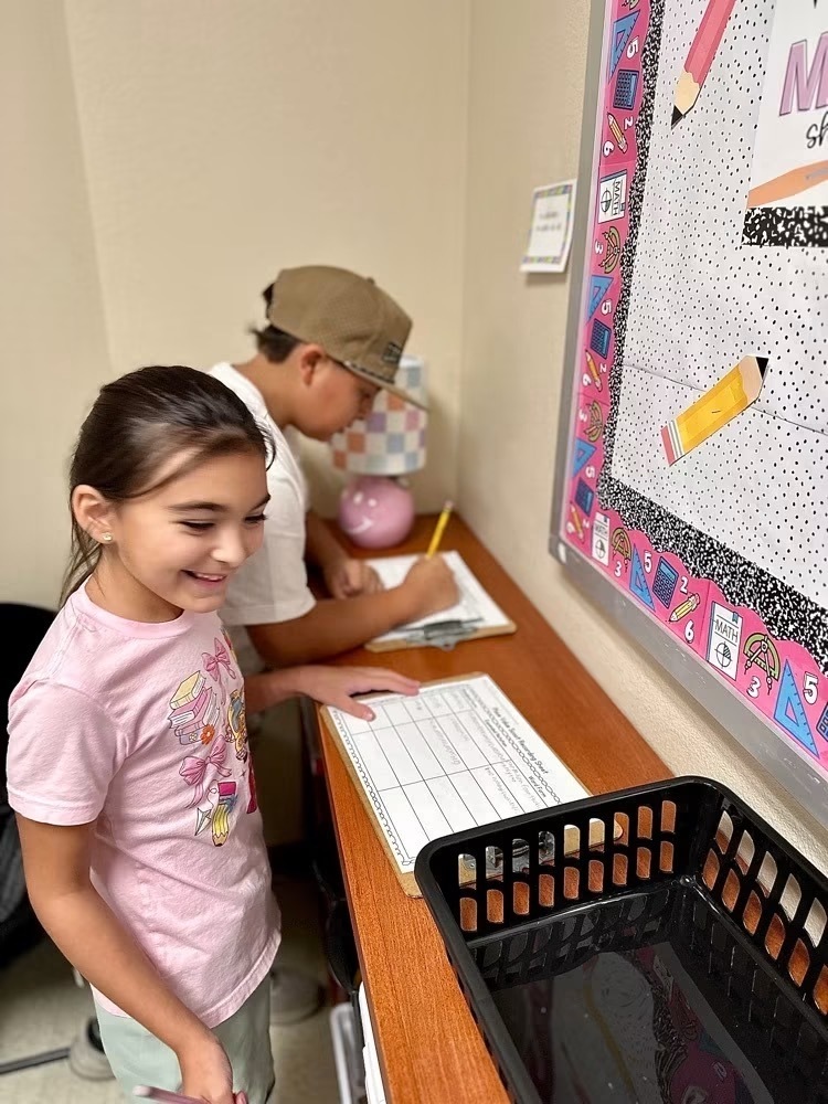 Two children, a girl in pink and a boy in brown, are standing by a table, with the boy writing.