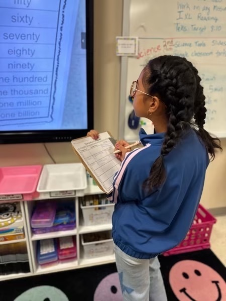A girl with braided hair writes on a notepad in a classroom. Behind her is a monitor displaying numbers.