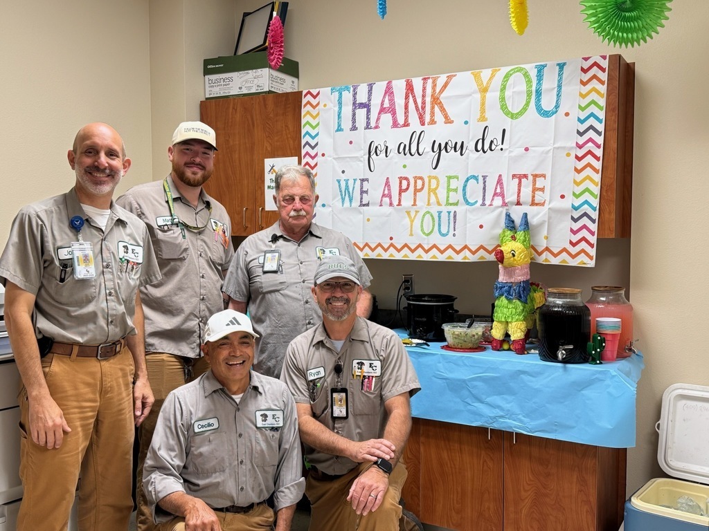 Five men in gray shirts and pants pose in front of a table with a thank-you banner.