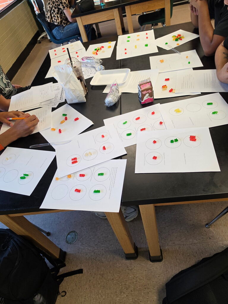 A classroom setting with students at a table, engaged in a learning activity involving colorful beads on paper.