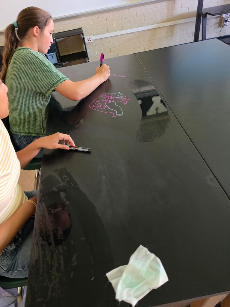 Two individuals seated at a black desk, drawing on a table's surface with pink and purple markers.