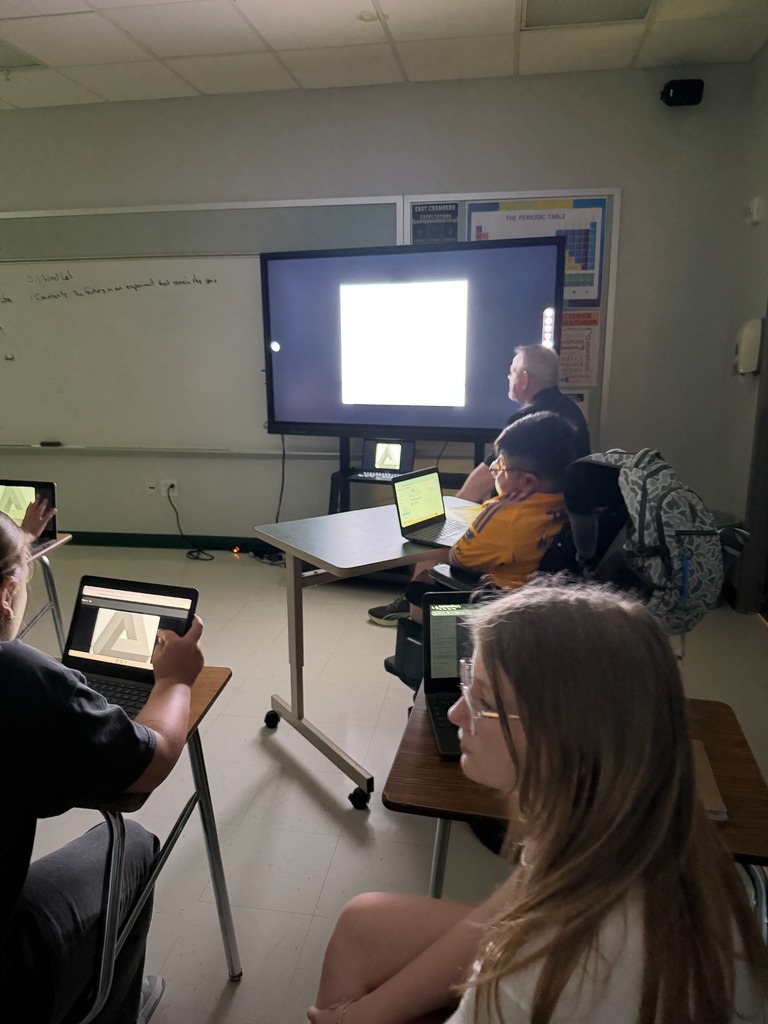 A classroom with students using laptops. A person at the front is projecting on a screen.
