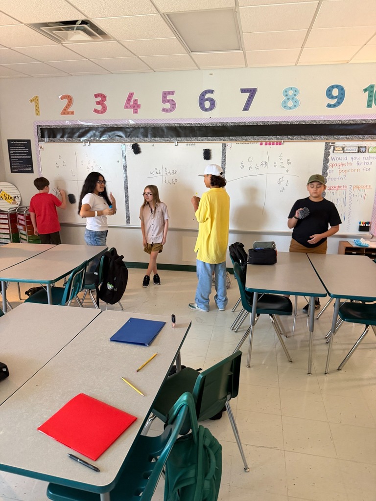 A classroom with tables and chairs. Five students are present. One student is writing on a whiteboard.