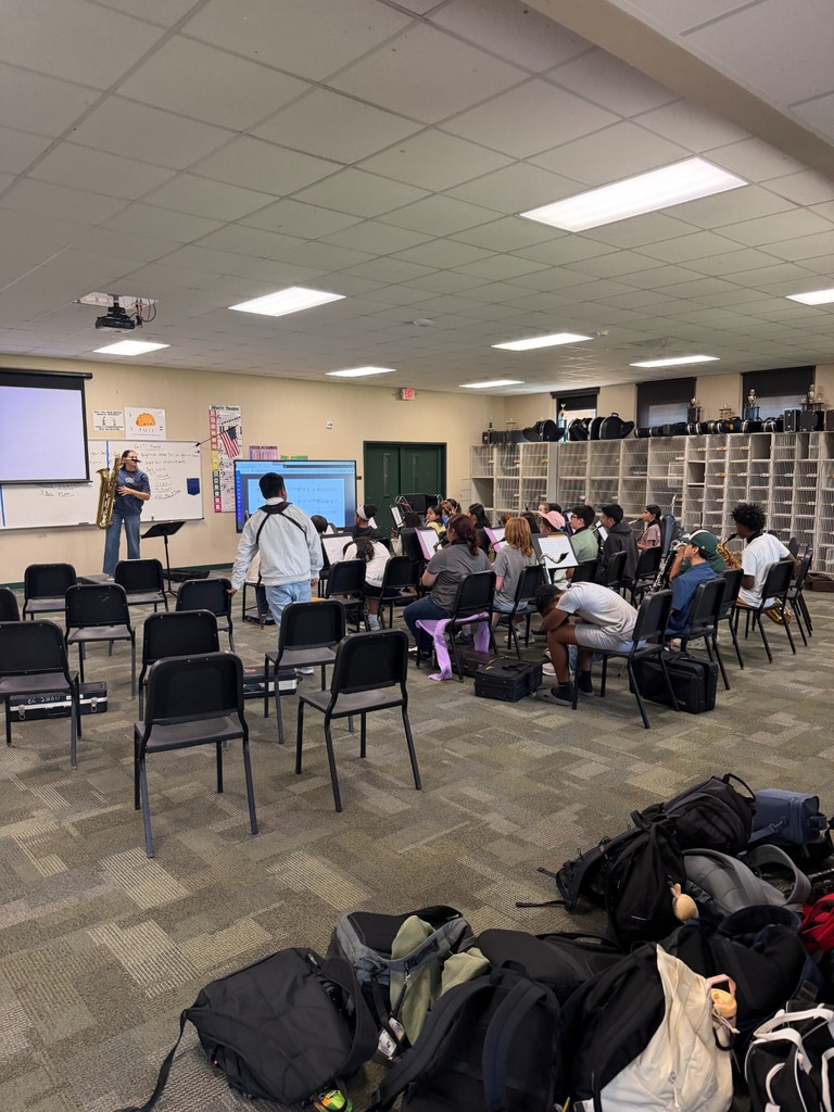 A classroom with chairs arranged in rows, a projector screen, a person standing at a podium, and many bags on the floor.