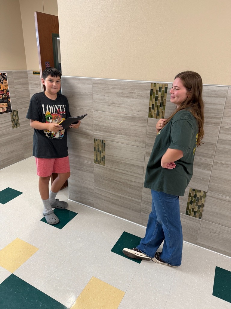 A young boy and a woman stand in a hallway, facing each other. The boy holds a tablet, and they appear to be conversing.