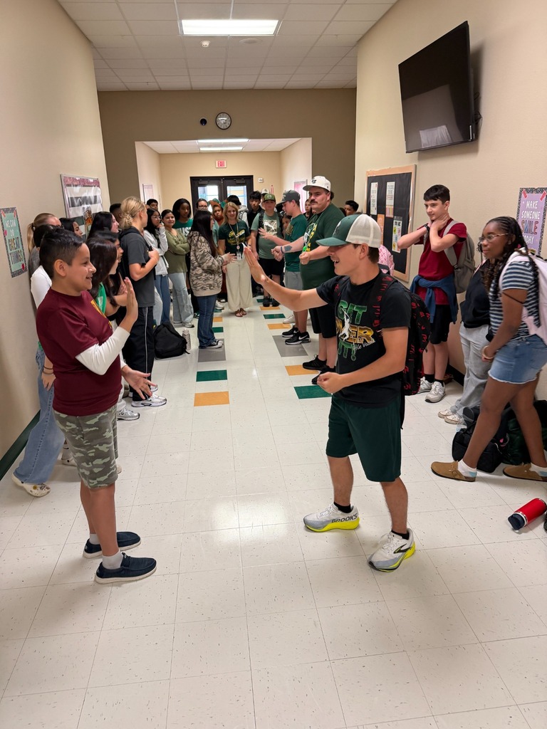People in a hallway with white floors, lined with a yellow and green checkered pattern, and a TV and clock on the wall.