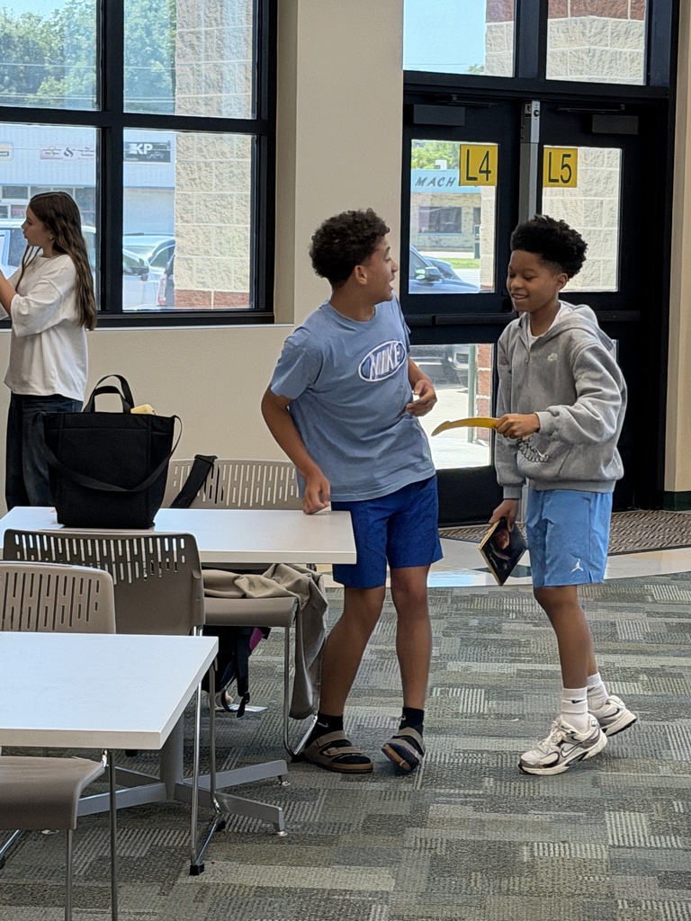 Two boys are standing near a table in a classroom. One holds a yellow object. A woman stands behind them.