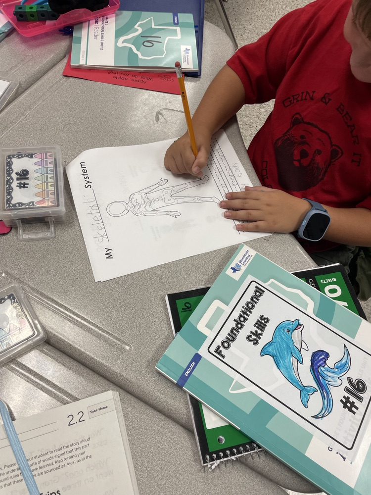 A child in a red shirt uses a pencil to draw on paper at a table. A green binder with "Foundational Skills" is in front of them.