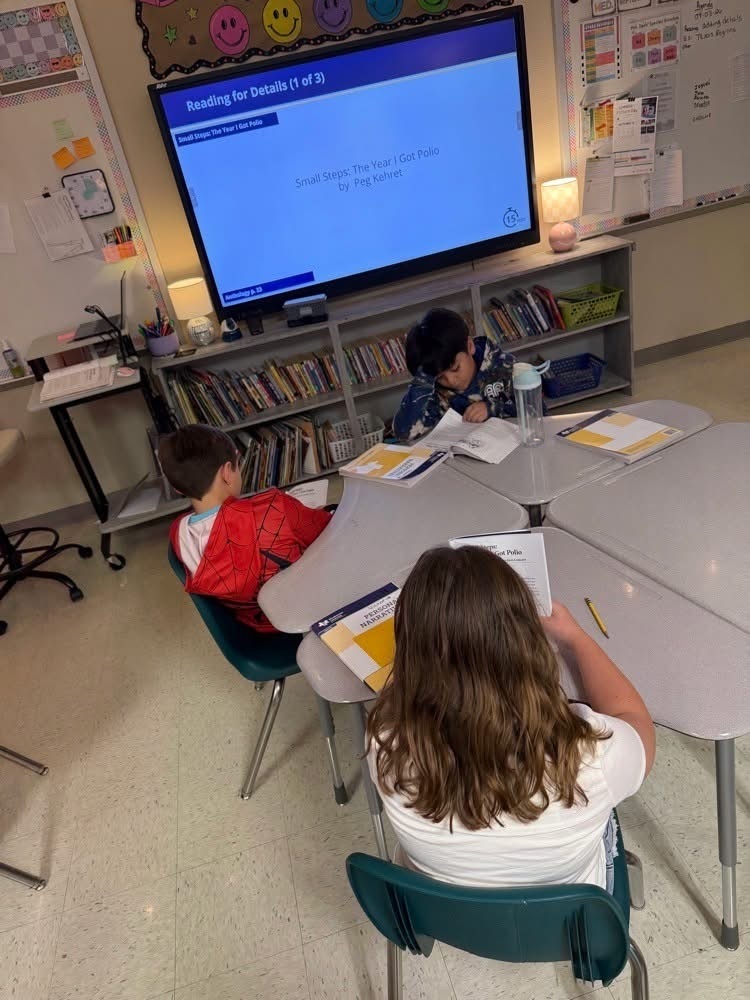 Three children in chairs at a table, reading. A monitor with a blue screen is on the wall behind them.