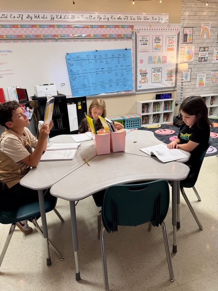 Three children seated at a round table, engaged in reading activities. Brightly colored containers and books are on the table.