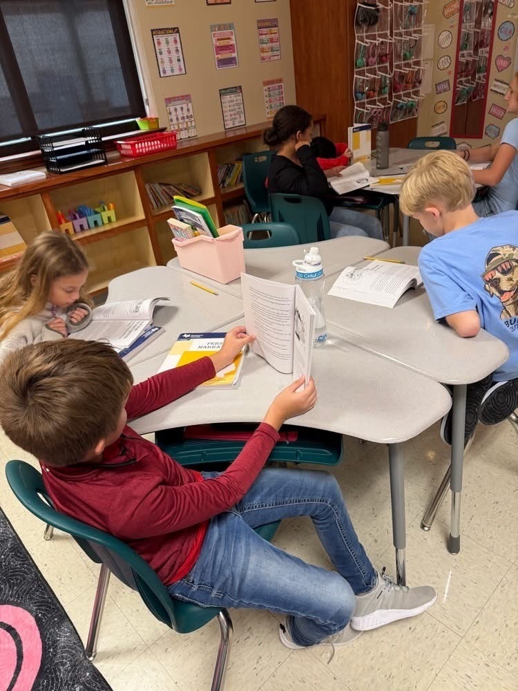 Four children seated at tables in a classroom, studying their books and papers. Shelves and a window are in the background.