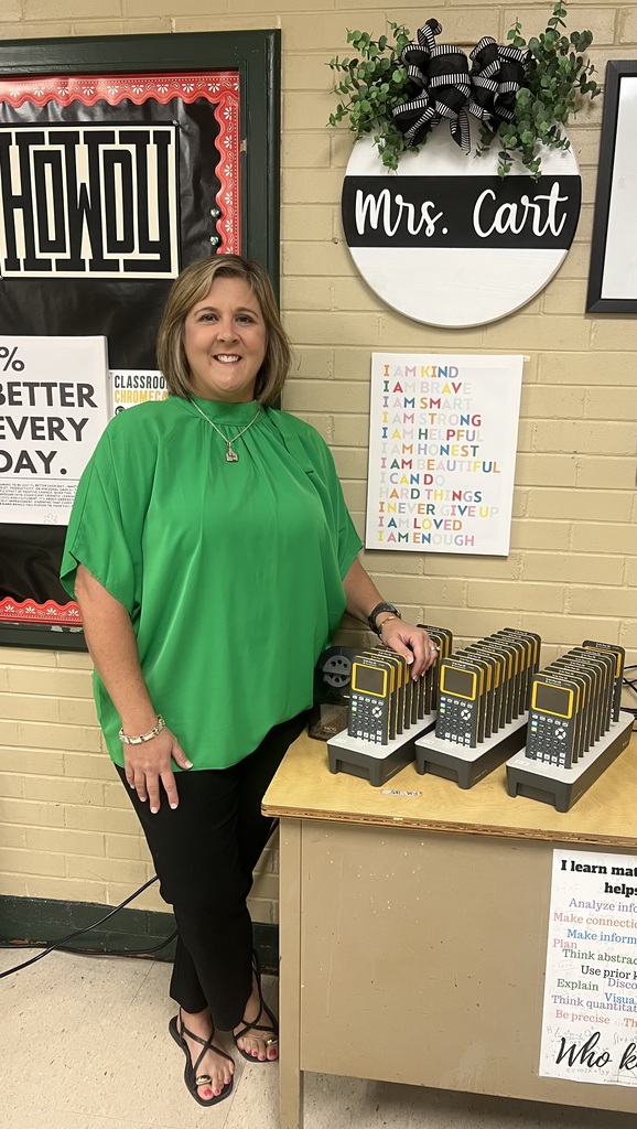 Woman standing at a table with calculators and a sign that reads "I learn math helps". Behind her, a wall with a sign saying "HOWDU" and a plant.
