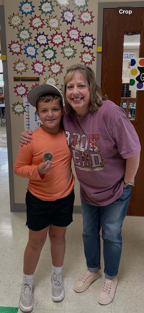 Woman in purple shirt with child in orange shirt, holding something green, smiling, in a classroom.