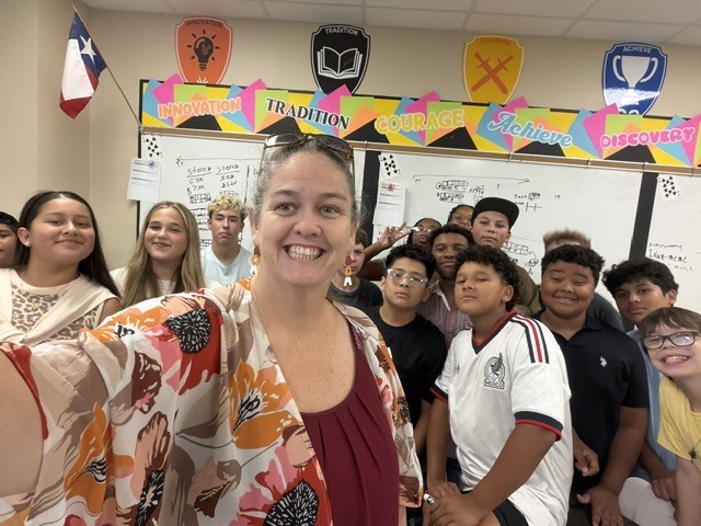 A woman smiles, taking a selfie in a classroom with a group of students in front of a whiteboard.