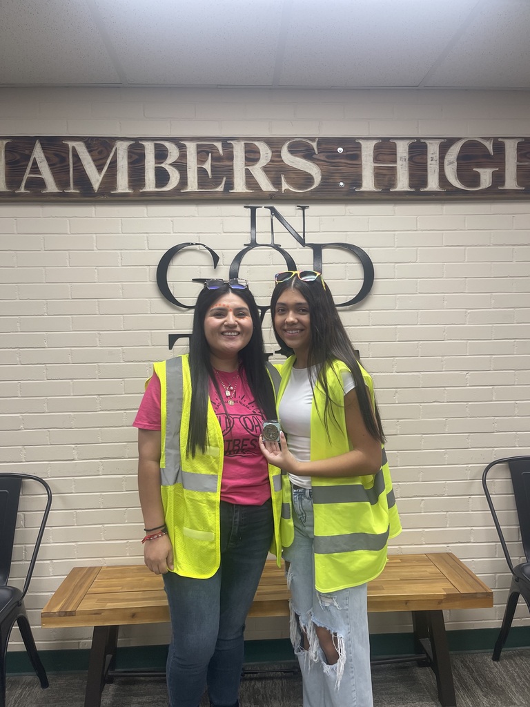 Two women in high-visibility vests smile in front of a sign. The sign reads "Chambers High in God."