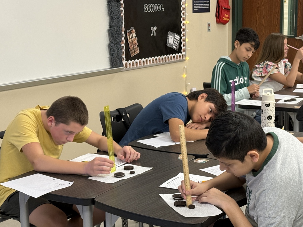 Four boys in a classroom. Each boy holds a stick and a pencil. The room has a white board and chairs.