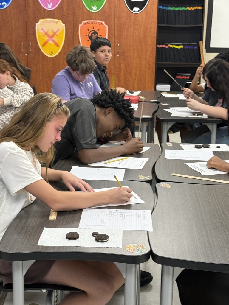 A group of students sit at desks and work on a science activity. One student leans forward.