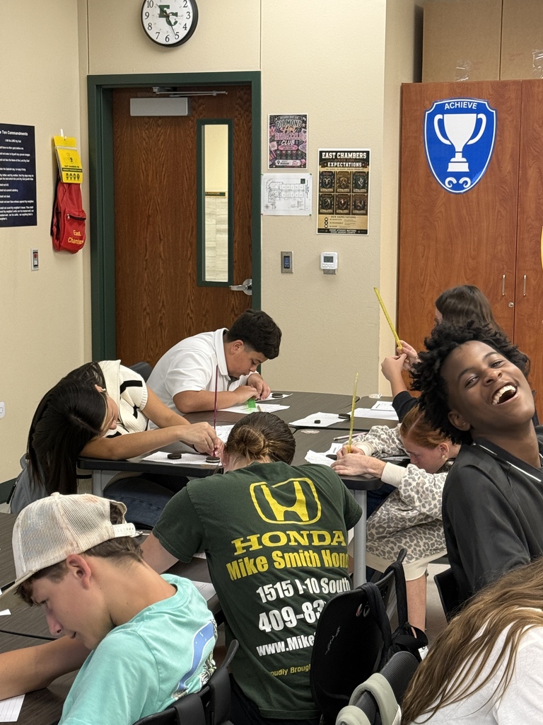 Students seated at tables in a classroom with a clock, a trophy, and a door in the background.