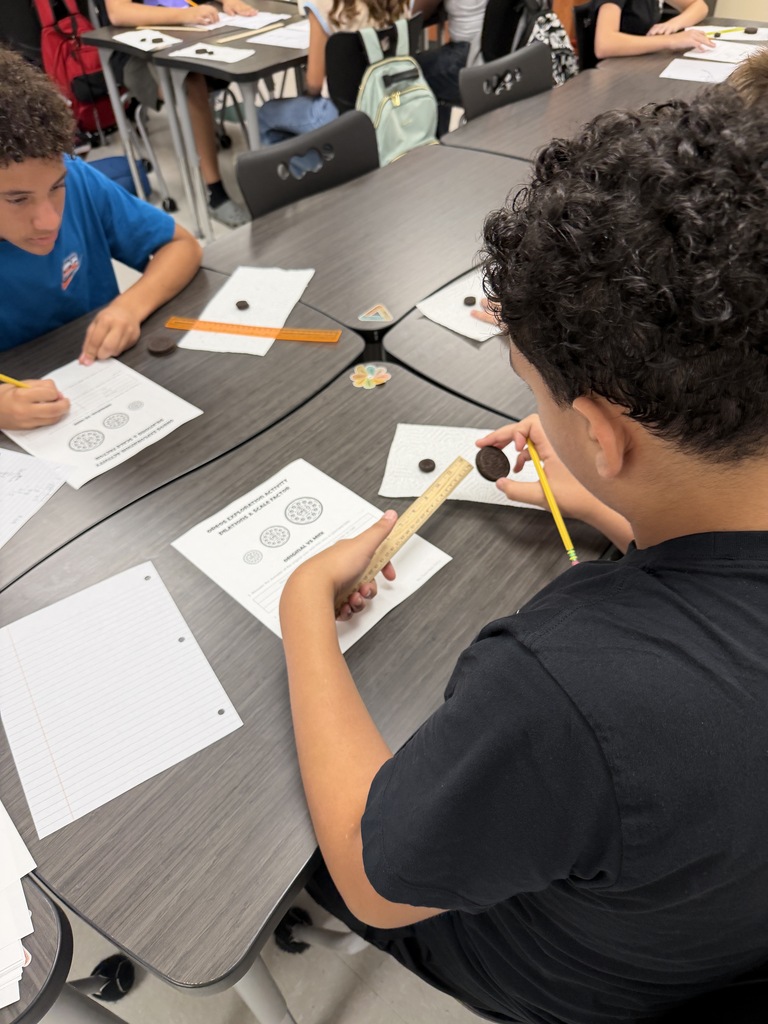 Students sit at desks, engaged in a lesson. One student uses a wooden stick to point at circles on a worksheet.