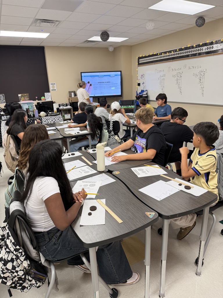 Students sitting at tables in a classroom with a teacher standing in front of a projector screen.