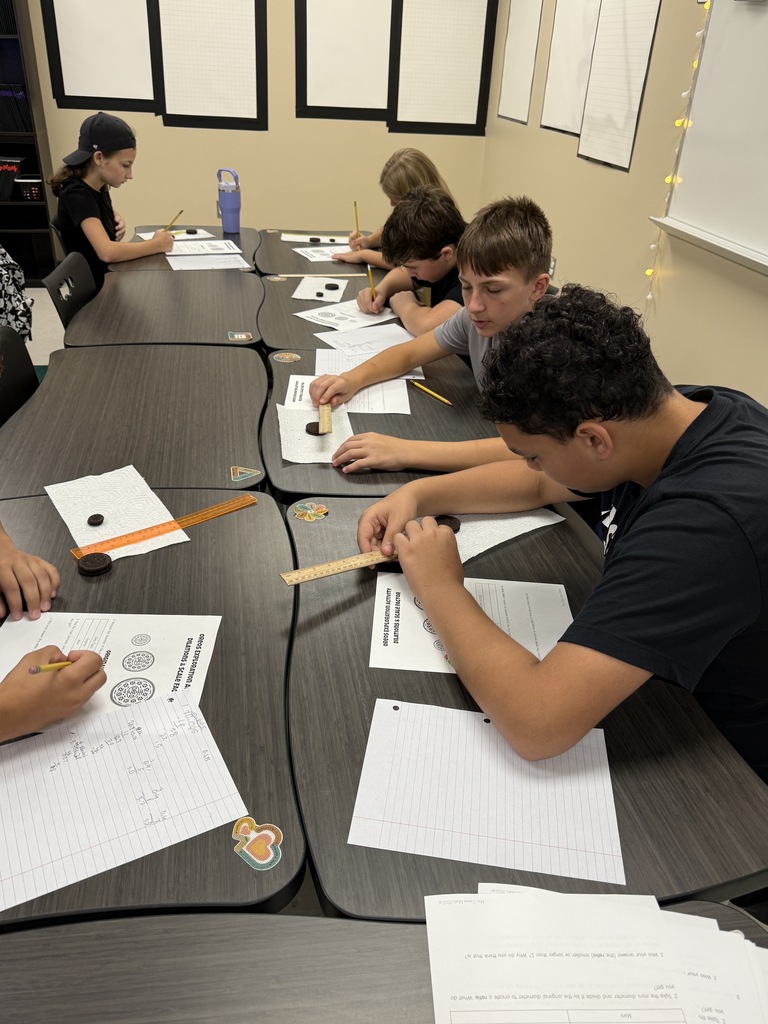 Students sit at tables and work on a hands-on activity with a wooden stick and paper.