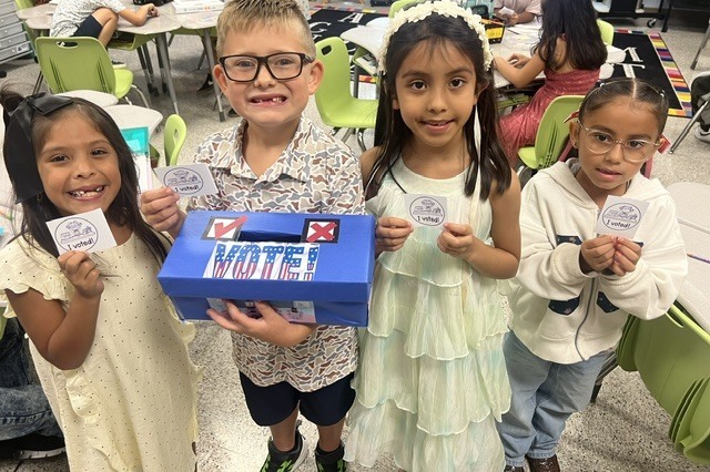 Four children, holding cards, stand in front of a blue box labeled "VOTE!" with a classroom in the background.