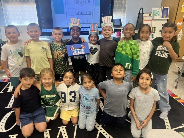 Several children in casual attire, some wearing hats, gather together in a classroom, smiling for a photo.