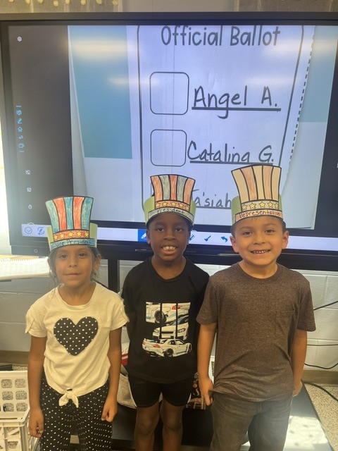 Three children stand next to a screen with "Official Ballot" and two names. They wear hats with stars.