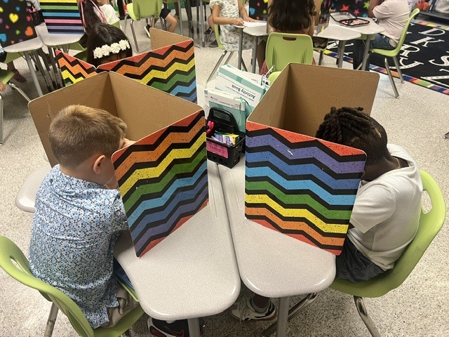 Two young children seated at a desk, each with a box-like divider between them, in a classroom.