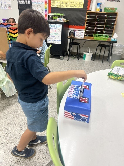 A boy in a blue shirt and shorts at a table inserts a card into a blue box.
