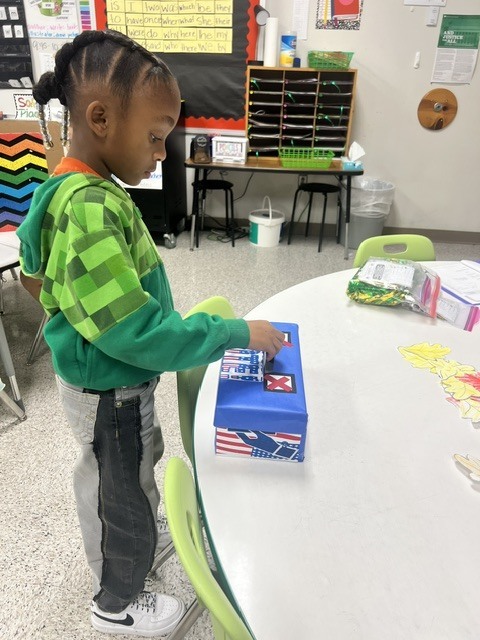 A young child in a green hoodie and sneakers stands at a table, opening a blue box.