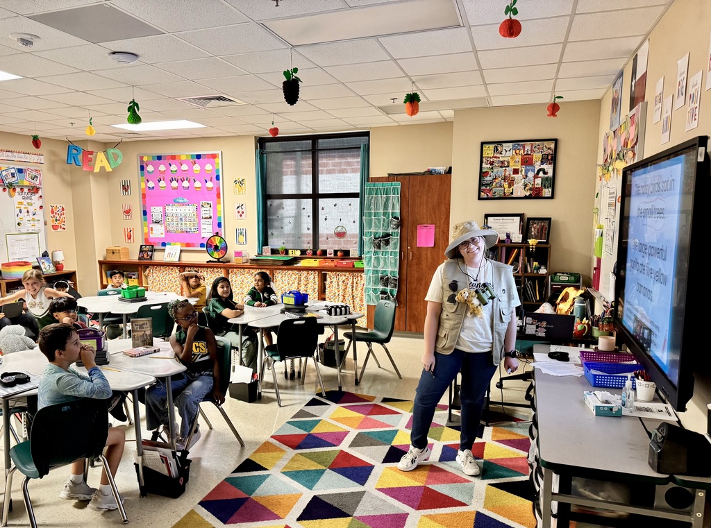 An adult stands in front of a classroom with students seated at desks. A monitor is on a stand next to a table.