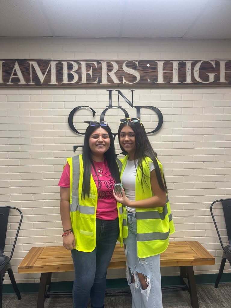 Two women in reflective vests stand next to a wooden bench. Behind them, a wall has "Chambers High In God" sign.
