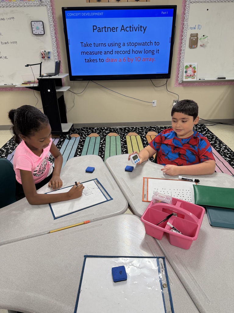 Two children, a girl and a boy, sit at a desk in a classroom, using a stopwatch and paper.