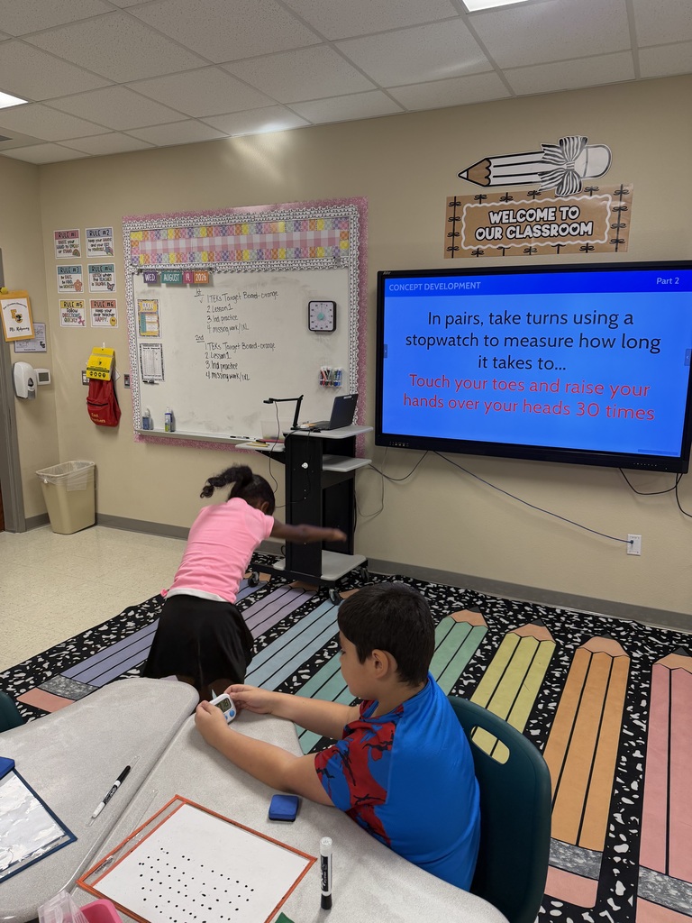 Two children seated at a desk in a classroom. A large screen displays a message. A whiteboard is on the wall.