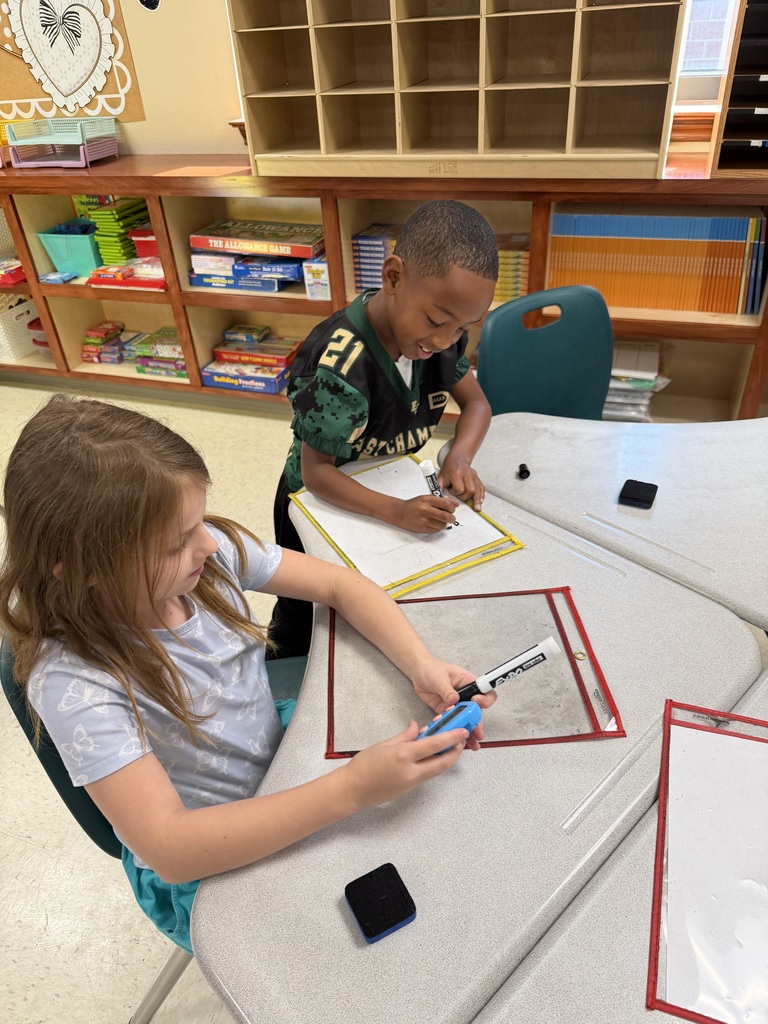 Two kids sit at a desk, with one writing and the other holding a blue pen.