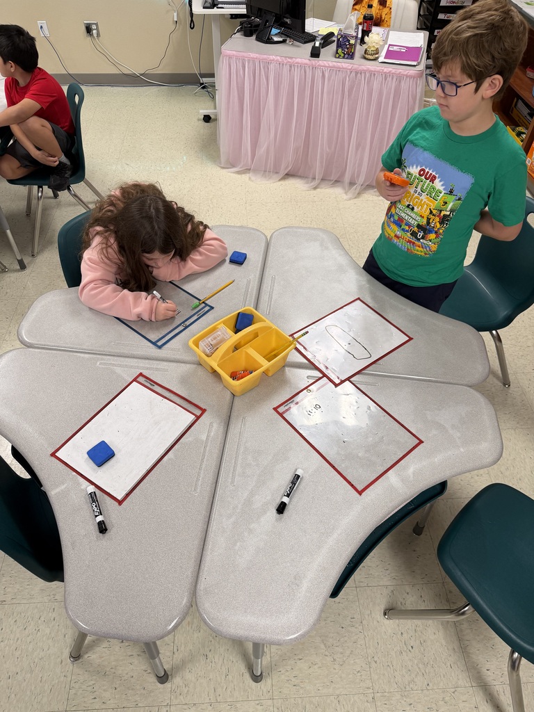 Two children sit at a table, one working on a piece of paper while the other stands nearby.