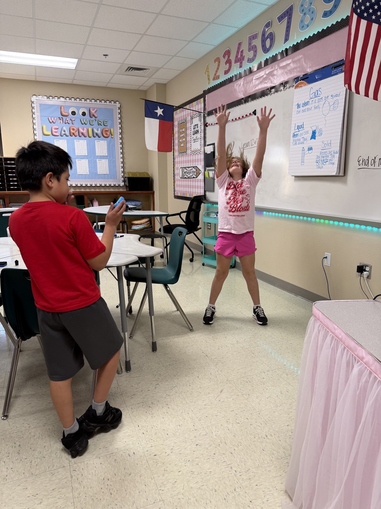 Two children playing with a cell phone in a classroom. The child in red is looking at a phone.