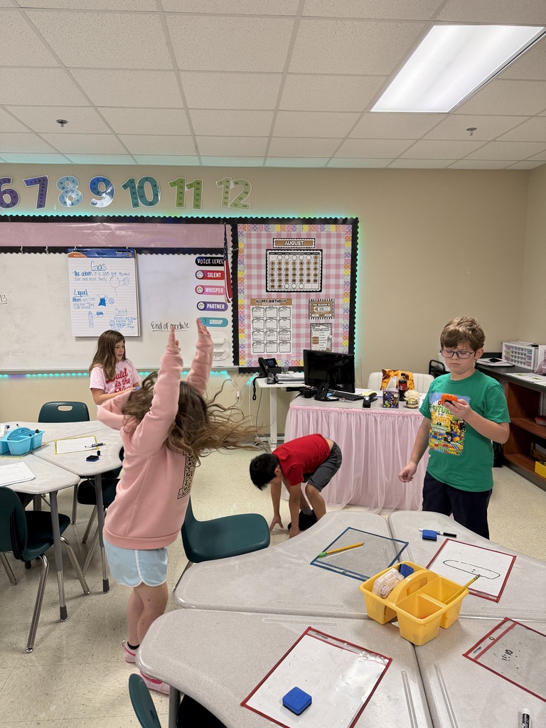 Classroom with four children. A girl stands with arms raised. Another bends over a table. Two boys stand next to a table with blue blocks.