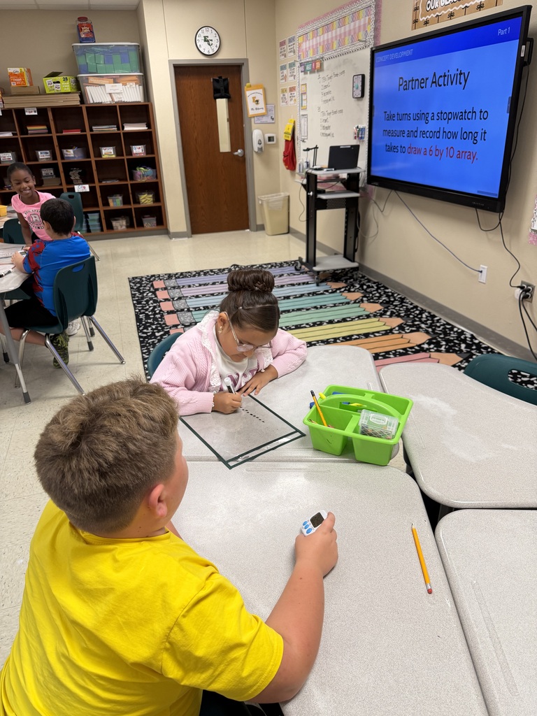 A classroom with children seated at tables. A boy in a yellow shirt holds a device. A girl in a pink sweater looks at a paper. A screen is on the wall.